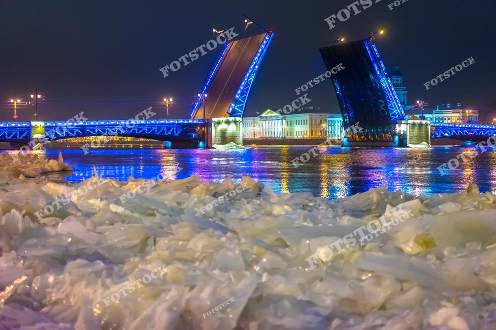 На фотографии запечатлён Дворцовый мост в Санкт-Петербурге, разведённый для прохода судов, украшенный яркой голубой подсветкой. Вода Невы покрыта ледяными глыбами, что добавляет ощущение зимнего холода и суровости пейзажу. На заднем плане видны исторические здания и их отражения, создающие живописную ночную панораму. Разведение мостов — один из символов ночной жизни Петербурга, привлекающий внимание туристов и жителей города.
